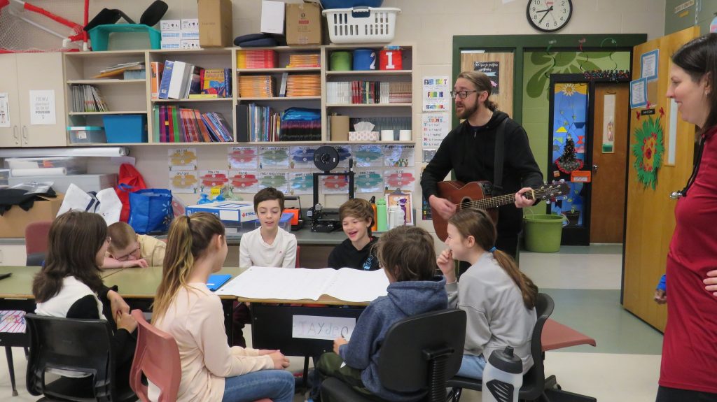 Un artiste joue de la guitare des élèves l'accompagnent au chant dans un contexte d'art et interdisciplinarité à l'école.
